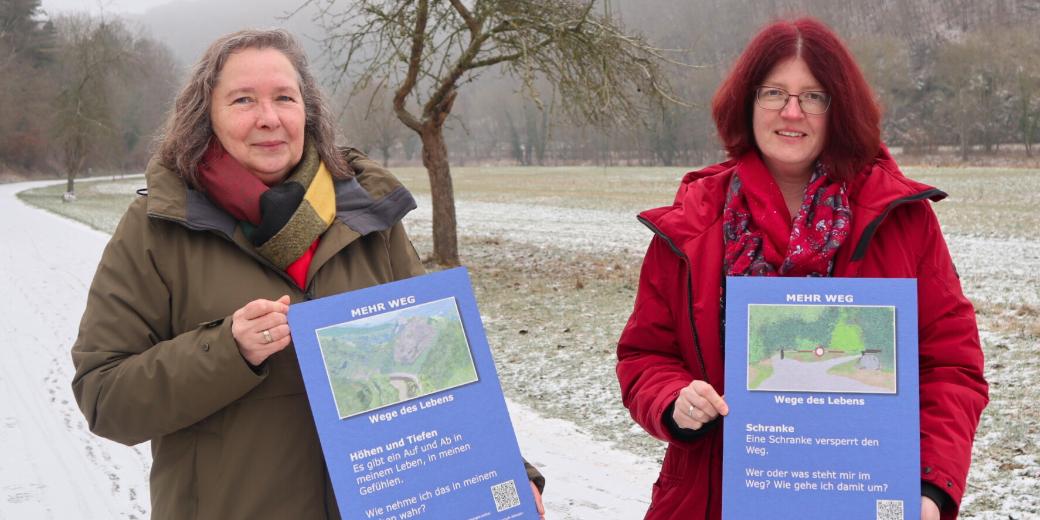 Zwei Frauen stehen auf einem Weg. Jede hält eine Bildtafel in der Hand.